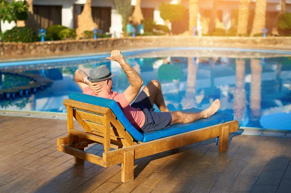 Relájese en la piscina de verano. Joven y exitoso hombre con sombrero acostado en una tumbona en el hotel en el fondo de la puesta del sol, concepto de tiempo para viajar — Foto de Stock