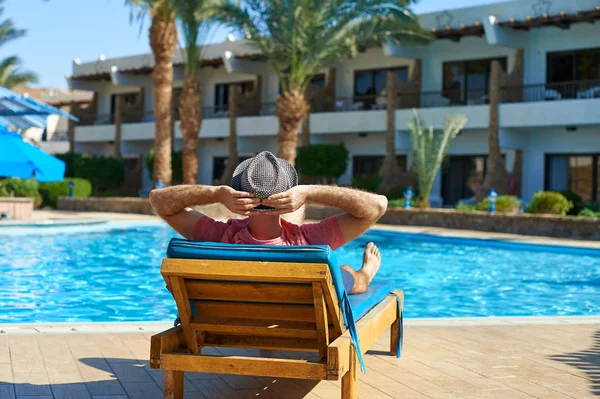 Relájese en la piscina de verano. Joven y exitoso hombre con sombrero acostado en una tumbona bronceado en el hotel en el fondo de la puesta del sol, concepto de tiempo para viajar — Foto de Stock