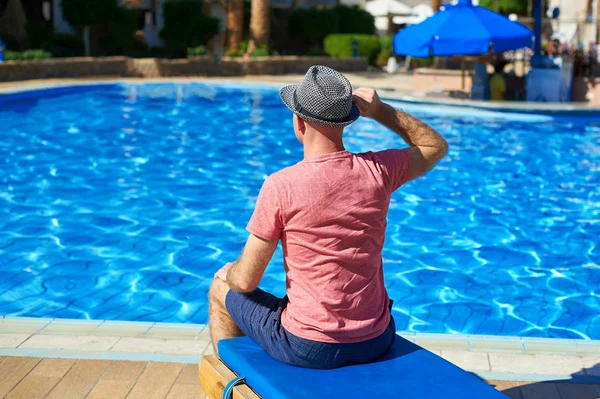 수영장 근처의 태양 안락에 앉아 모자에 있는 젊은 남자, 여행 하는 개념 시간. 여름에는 수영장에서 휴식을 취하십시오. — 스톡 사진