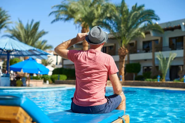 Молодой человек в шляпе сидит на шезлонге возле бассейна, концептуальное время для путешествий. Отдых в бассейне летом в Египте — стоковое фото