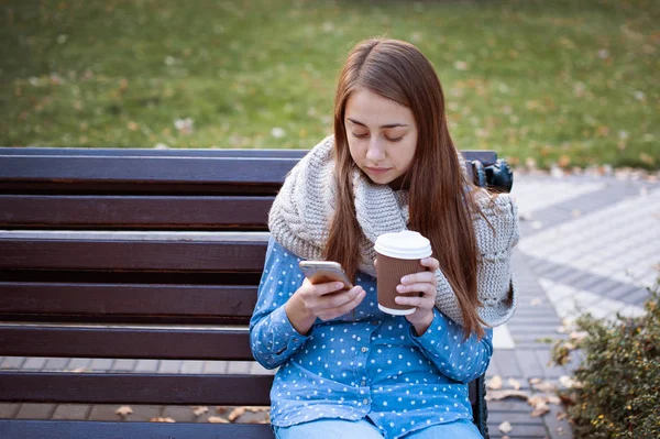 Chica joven y atractiva sentada en un banco en el parque de otoño y beber café — Foto de Stock