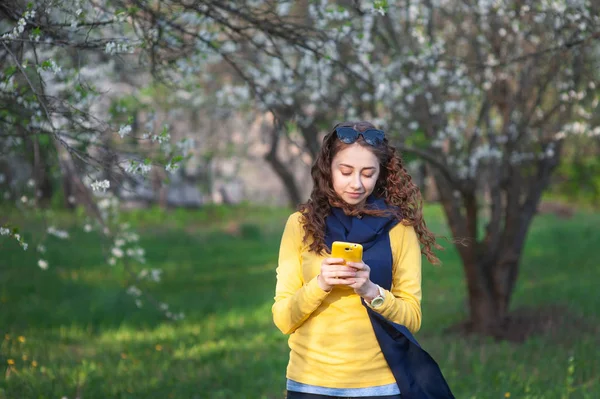 Bir çiçeklenme bahçesinde ayakta genç güleryüzlü kadın ve cep telefonu yazıyor. Kiraz çiçeklenme. Güzel kadının portresi — Stok fotoğraf