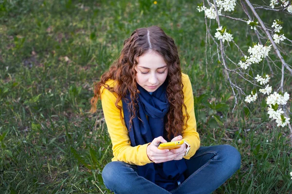 Genç güleryüzlü kadın bir çiçeklenme bahçede oturan ve cep telefonu yazıyor. Kiraz çiçeklenme. Güzel kız portresi — Stok fotoğraf