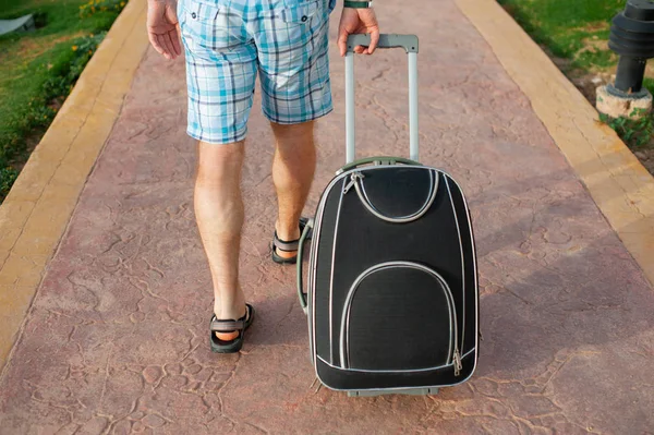 Молодий чоловік тягне валізу на дорогу в літньому парку. Мандрівний хлопець у розумних шортах виходить зі своїм багажем — стокове фото