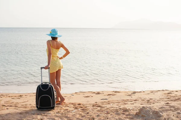 Молодая красивая женщина в желтом платье и шляпе с большим чемоданом на тропическом пляже. Девушка смотрит на море — стоковое фото