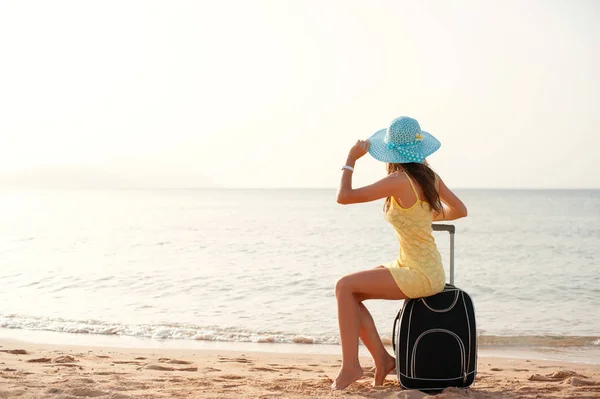 Seyahat tatil ve insanlar kavramı-kadın turist bavul ve şapka üzerinde deniz yakınında oturan ve güneş izlerken ve göstermek thumbs up — Stok fotoğraf