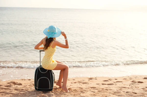 Seyahat tatil ve insanlar kavramı-kadın turist bavul ve şapka üzerinde deniz yakınında oturan ve güneş izlerken ve göstermek thumbs up — Stok fotoğraf