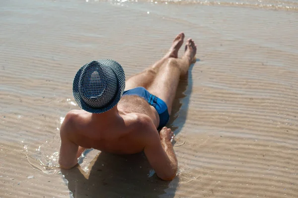 Elegante jovem homem modelo deitado na areia da praia usando chapéu de verão hipster desfrutando de férias de verão perto do mar — Fotografia de Stock