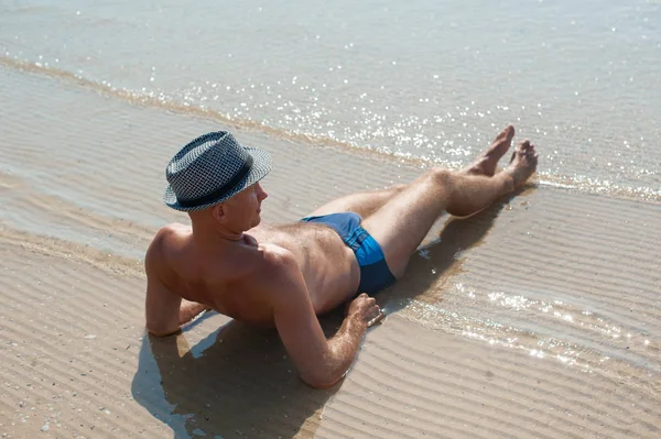 时尚年轻男模特男子躺在沙滩上,戴着时髦的夏帽享受海边的暑假 — 图库照片
