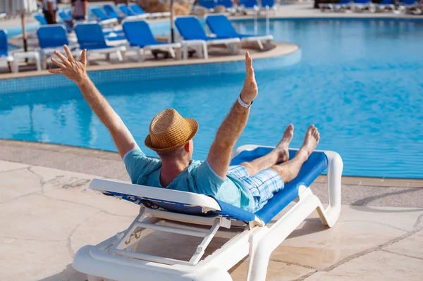 Hombre en sombrero tomando el sol en una tumbona junto a la piscina de verano. Concepto de viaje y descanso — Foto de Stock