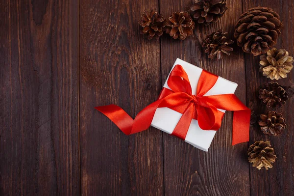 Kartki świąteczne pozdrowienia. Białe pudełko z czerwoną wstążką i guzki na drewnianym tle z miejscem na tekst — Zdjęcie stockowe
