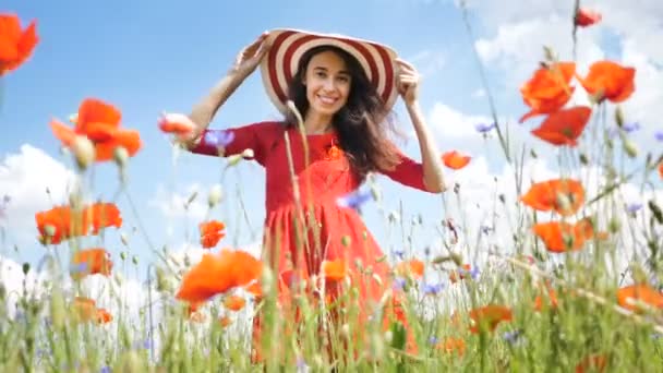 赤いドレスと大きな帽子で幸せな若い女性が自然を楽しんでいます。美容室屋外はケシ畑を歩いています。自由の概念。空と太陽の上の美少女 — ストック動画