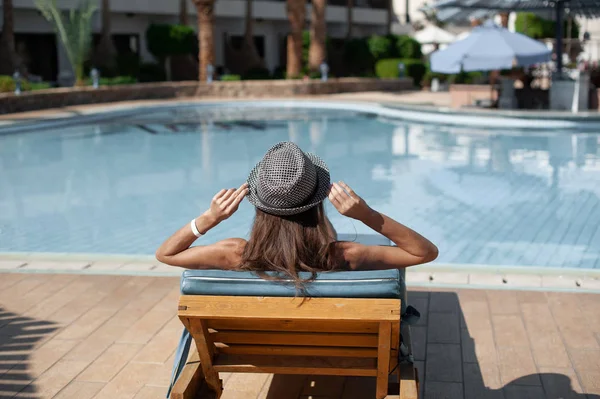 Jeune belle femme assise sur des chaises longues près de la piscine à l'hôtel en chapeau — Photo