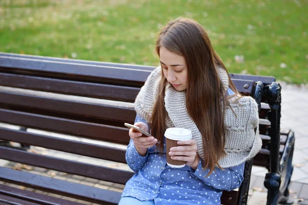 Menina jovem e atraente sentado em um banco no parque de outono beber café e falar no smartphone móvel — Fotografia de Stock