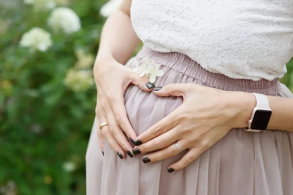 Donna fa una forma di cuore con le mani sopra la vita in espansione, il suo ombelico centrato. Fiori e piante sfondo — Foto Stock