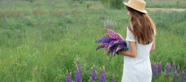 Lupine Field Pankartı 'nda güzel bir kadın. Beyaz elbiseli ve şapkalı bir buket lupin tutan tatlı bir kız. Yaz zamanı. Arka plan