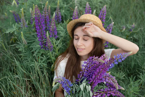 Mulher romântica bonita com buquê de tremoços sorri alegremente em vestido branco e chapéu senta-se no campo de flores lupinas roxas. Foco seletivo suave — Fotografia de Stock