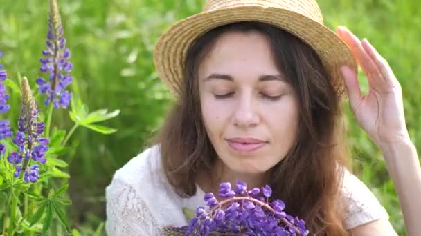 Hermosa mujer romántica con ramo de altramuces sonríe alegremente en vestido blanco y sombrero se sienta en el campo de flores de altramuces púrpura. Enfoque selectivo suave — Vídeo de stock