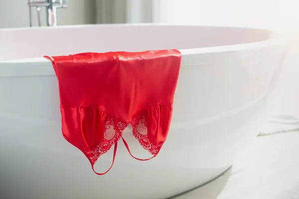Luxurious satin nightgown in red hung on the bathtub in the bathroom in the morning.