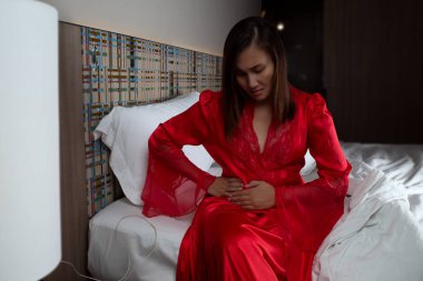 Have a pain under right waist. A woman in satin nightgown and red robes in pain holding his stomach and rib pain on right side in the bedroom at night. clipart