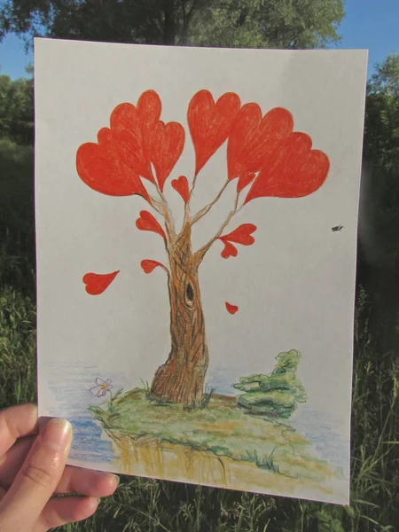 Een hand houdt een tekening van een fantastische boom met rode harten, waarop een bug zit, in het licht van de rijzende zon. — Stockfoto