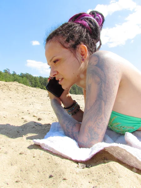 Egy lány dreadlocks, tetoválások és a piercing egy napsütötte tengerparton fekszik a homokon, és komolyan beszél a telefon. — Stock Fotó