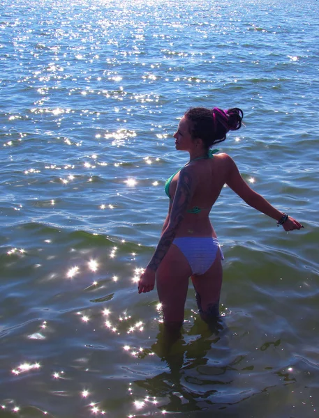 Una chica bronceada con rastas púrpuras, tatuajes y un piercing está en el agua contra la superficie del mar . — Foto de Stock