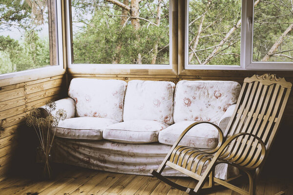 A wooden rocking chair and a sofa on the porch or terrace with a view of the forest. Tinted image