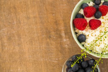 Taze Ahududu ve yaban mersini rustik ahşap arka plan üzerinde olgun çilek ile sağlıklı kahvaltı için bir kase süzme peynir. Düz yatıyordu.