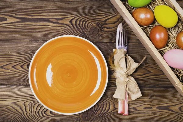 Easter table setting with empty orange plate and cutlery and multicolored eggs in a box on a wooden table. Holidays background.