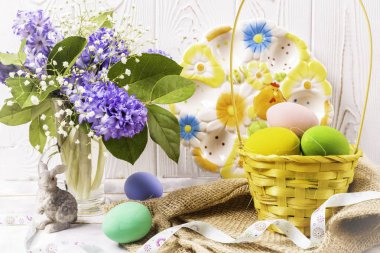 Bir hasır sepeti ve bir tavşan heykelcik ve bir vazo ve beyaz ahşap masa üzerinde güzel bir seramik plaka bir bahar buket Paskalya renkli yumurta