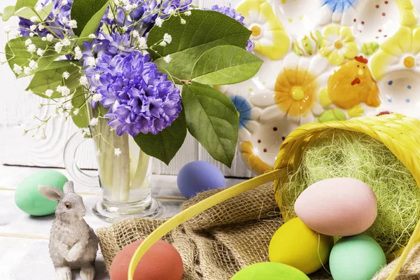 Œufs colorés de Pâques dans un panier en osier et une figurine de lapin et un bouquet de printemps dans un vase et une belle assiette en céramique sur une table en bois blanc — Photo