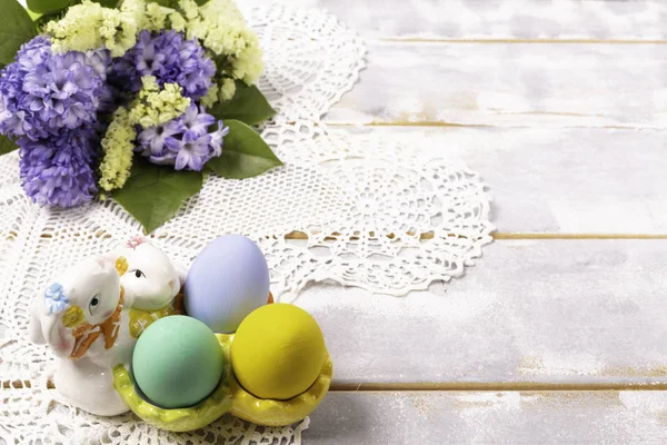 Œufs de Pâques colorés dans un stand de lapin en céramique et un bouquet de printemps avec des fleurs lilas et jaunes sur une belle serviette tricotée sur une table en bois blanc — Photo