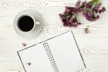 Beyaz ahşap bir masa üzerinde üstten görünüm bir günlük ya da defter ve kahve ve mor bir çiçek. Düz tasarım.