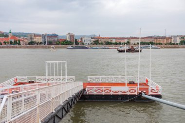 Budapeşte 'deki Tuna Nehri' nin manzarasına göre limanda yüzen bir rıhtım ve denizde yüzen tekneler var. Arka planda, tarihi ve modern binalar bulutlu bir gökyüzünün altında nehir kıyısına dizilir.