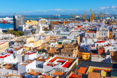 Hava görüntüsü yoğun bir Cadiz şehri, beyaz binalar ve liman manzarası gösteriyor. Turnalar ve bir köprü arka planda görünür, deniz ufka kadar uzanıyor.