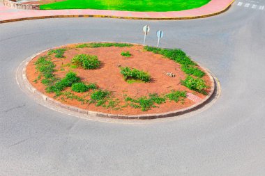 Nadir bitki örtüsü ve yol işaretleri olan merkezi bir dikim yatağı içeren dolambaçlı bir yol. Çalılarla döşenmiş dairesel trafik adası asfalt yolların kesişiminde duruyor.