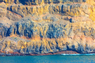 Yükselen kaya oluşumu sakin okyanustan dramatik bir şekilde yükselir. Uçurumun engebeli yüzeyi, altında su olan farklı katmanlar ve dokular gösteriyor. Sert ve pürüzlü yüzeyleri olan desenli jeolojik katmanlar.