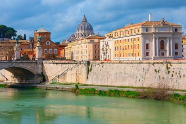 Tiber Nehri Roma 'daki Ponte Vittorio Emanuele II taş köprüsünün altından akar. Tarihi binalar bulutlu gökyüzünün altında nehir kıyısında yer alır. St. Peter Bazilikası 'nın kubbesi uzaktan görülebilir.