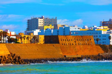 Okyanus kıyısı boyunca Cadiz 'deki Baluarte de San Roque' da, tarihi taş duvarlar dalgalara bakıyor. Kalenin arkasındaki modern binalar ve palmiye ağaçları Endülüs kıyı kenti yaşamını harmanlıyor.