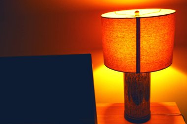 Turuncu abajurlu parlayan masa lambasının yer aldığı davet edici sahne, odada ortam ışığını yansıtıyor. Derinlik ve rahat atmosfer yaratıyor. Lambanın içindeki sıcaklığın hafifçe aydınlatması.