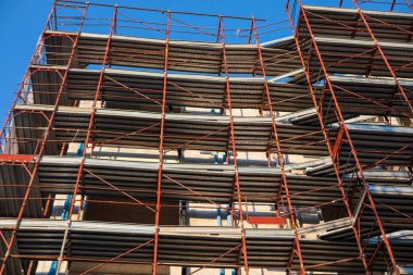 Metal iskelet iskeletleri yapım aşamasındaki çok katlı bir binayı çevreleyerek geometrik çizgiler ve platformlar oluşturur. Yapı gökyüzüne karşı yükseliyor, kentsel büyüme ve gelişimi simgeliyor