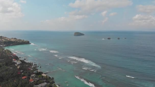 Filmagem aérea: Voando acima da ilha tropical litoral de Bali, aldeia local e resorts em direção a um oceano com ondas, palmas, areia cinza e falésias. 4K — Vídeo de Stock