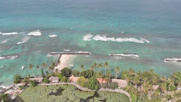 Imágenes aéreas: Volando por encima de la isla tropical de la costa de Bali, pueblo local y resorts hacia un océano con olas, palmeras, arena gris y acantilados. 4K — Vídeos de Stock