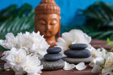 Gri Zen taşlarından piramitler, güzel taze beyaz çiçekler yeşil yapraklar ve ahşap arka planda Buda heykeli. Uyum kavramı, denge ve meditasyon, spa, masaj, rahatlama