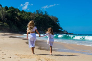Beyaz elbiseli sevimli kızıyla genç ve güzel bir anne sahile yakın kumsalda yürüyor.