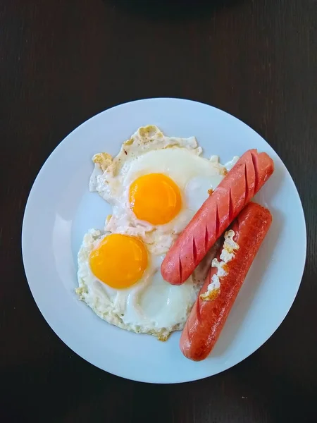 白い皿の上に卵2個とソーセージ2個の目玉焼きが木製のテーブルで朝食に出される — ストック写真