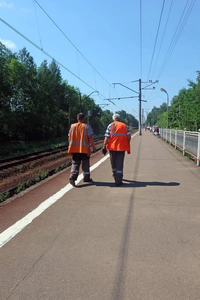 Dwóch Pracowników Pomarańczowych Kamizelkach Spaceruje Peronie Kolejowym Rzucając Cień Asfalt — Zdjęcie stockowe