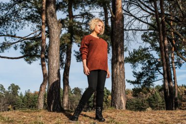 Sonbahar Park pines yakın duran sarışın kadın. Pişmiş toprak jumper ve açık havada etkinlik için ormandaki siyah kot kıvırcık saçlı kadın. Kavram - spor aktivitesi açık havada.