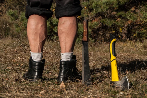 Strong legs of a man, standing near machete, stuck in the ground. Beautiful masculine legs with calf muscles, rear view. Concept - active lifestyle.